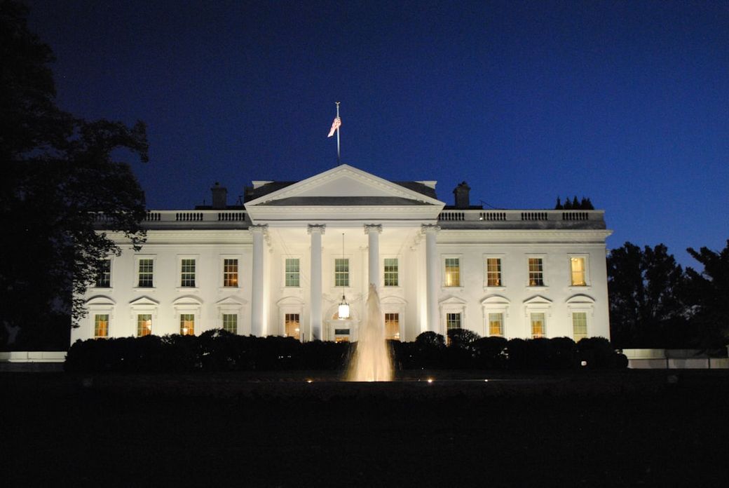 The image captures the exterior of the White House illuminated at dusk. The structure is framed by a blue sky and vibrant landscaping, showcasing the iconic architecture of the U.S. presidential residence. — Credit: Photo by Tabrez Syed on Unsplash