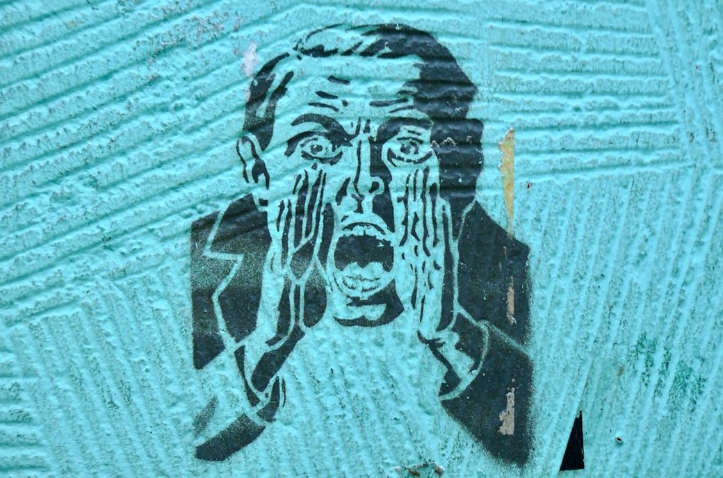 The image features a stencil graffiti art depicting a man screaming with his hands cupped around his mouth. The background is a textured turquoise surface that enhances the boldness of the artwork. — Credit: Photo by Bekky Bekks on Unsplash