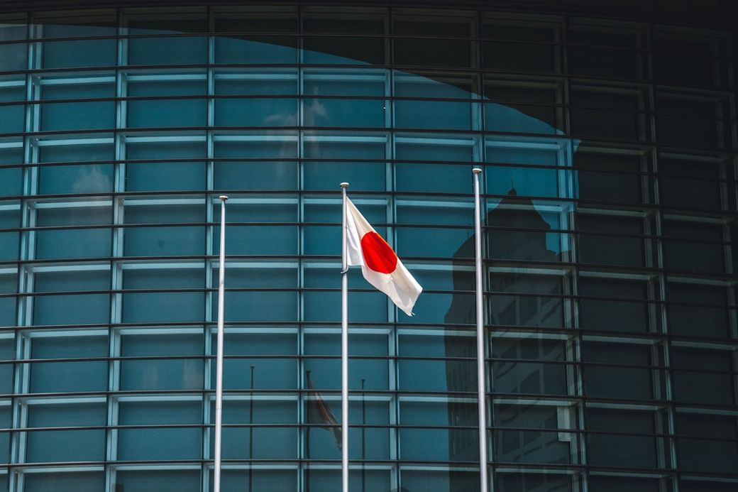 The image features the Japanese flag prominently displayed in front of a modern glass building. The flag's iconic red circle stands out against the sleek blue-tinted glass facade. — Credit: Photo by Roméo A. on Unsplash