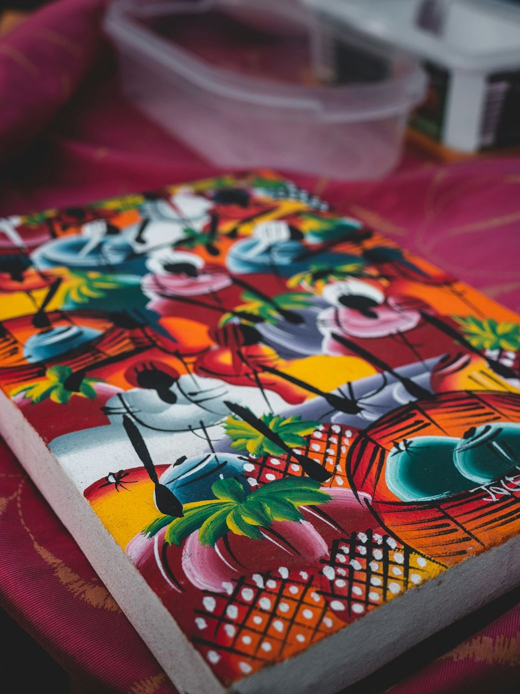 The image features a vibrant, colorful piece of artwork showcasing abstract figures and tropical elements. The painting is situated on a textured surface, with a backdrop that includes a softly patterned fabric. — Credit: Photo by Jonathan Kemper on Unsplash