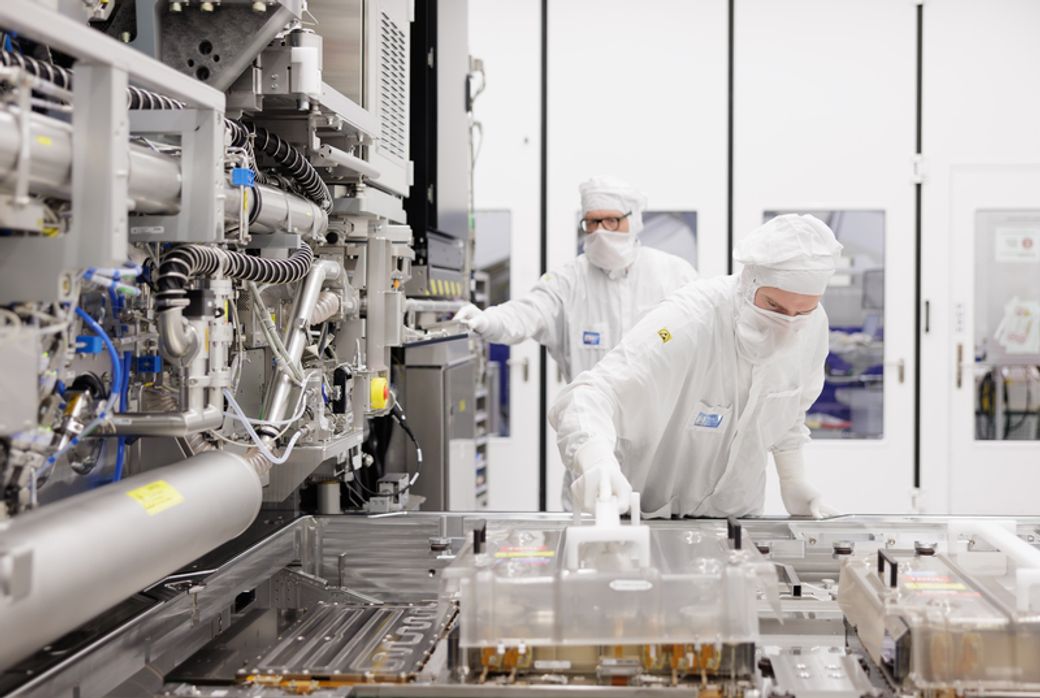 Two technicians in white protective suits are working in a cleanroom environment, focusing on equipment related to semiconductor manufacturing. The environment is sterile, designed to minimize contamination during production processes.