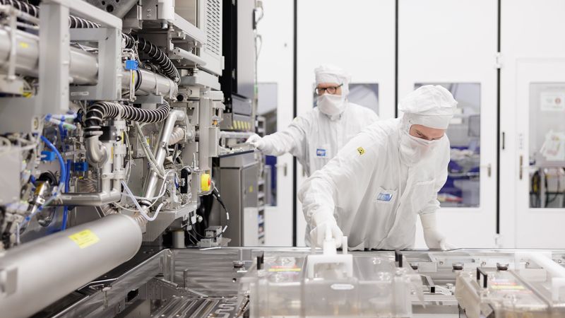 Two technicians in white protective suits are working in a cleanroom environment, focusing on equipment related to semiconductor manufacturing. The environment is sterile, designed to minimize contamination during production processes.