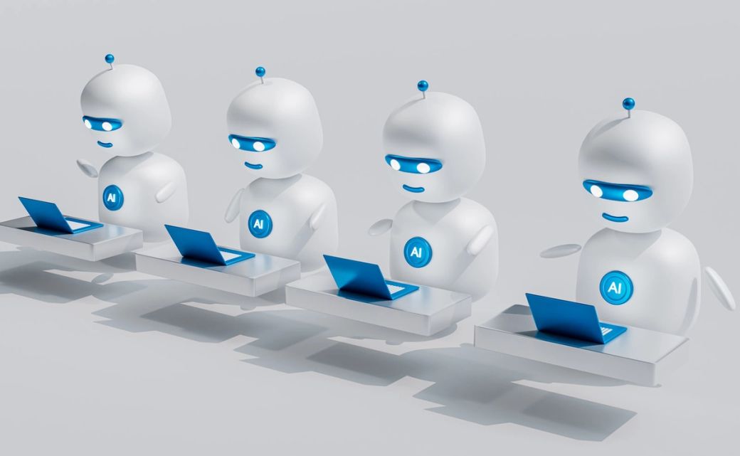 The image features four stylized robot figures seated at desks, each using a laptop. The robots are designed with smooth, rounded features and display blue accents that suggest a connection to artificial intelligence. — Credit: Photo by Mohamed Nohassi / Unsplash