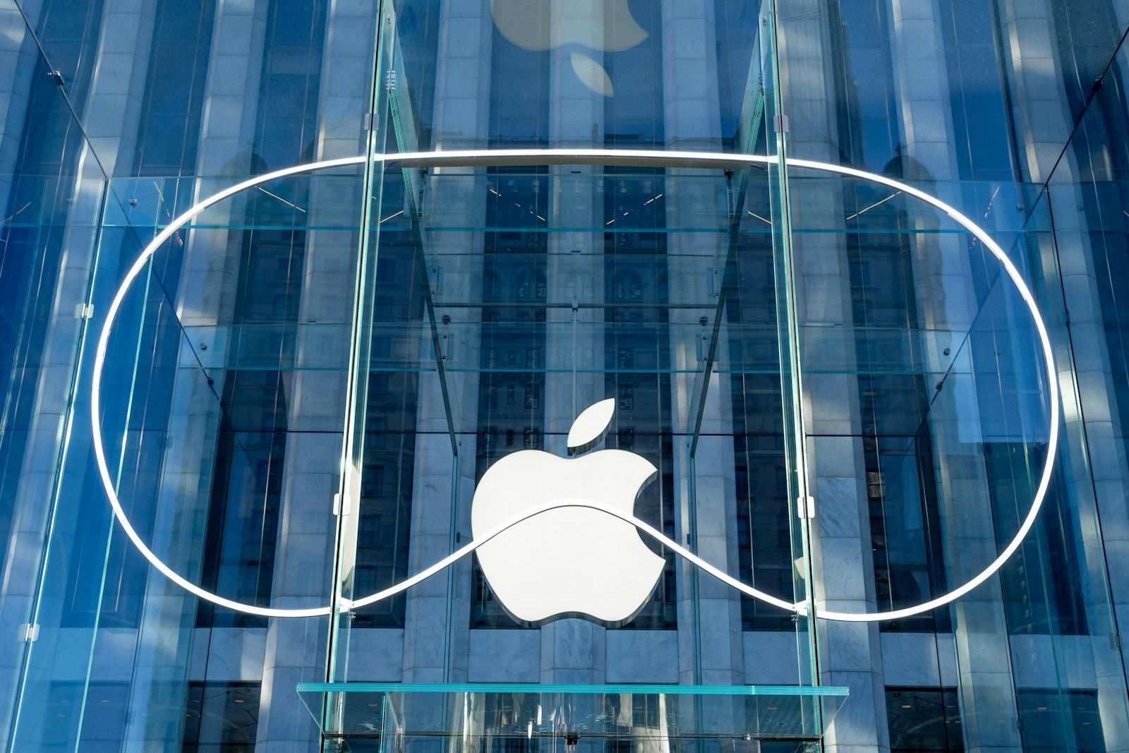 The image shows the exterior of an Apple retail store featuring a large, iconic Apple logo against a glass facade. The modern architectural design highlights the transparency and sleekness of the building's structure.
