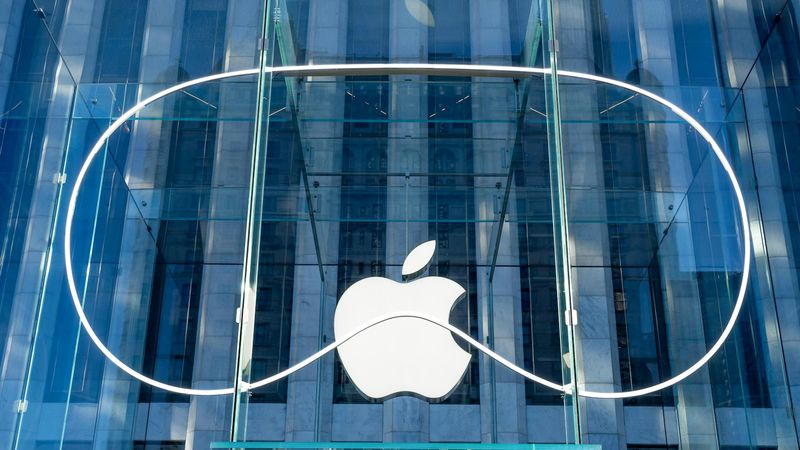 The image shows the exterior of an Apple retail store featuring a large, iconic Apple logo against a glass facade. The modern architectural design highlights the transparency and sleekness of the building's structure.