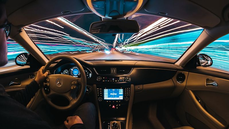 An interior shot of a car in motion, showcasing a driver actively steering the vehicle with blurred streaks of light passing by outside, indicating high speed. The dashboard is illuminated with various controls and displays.