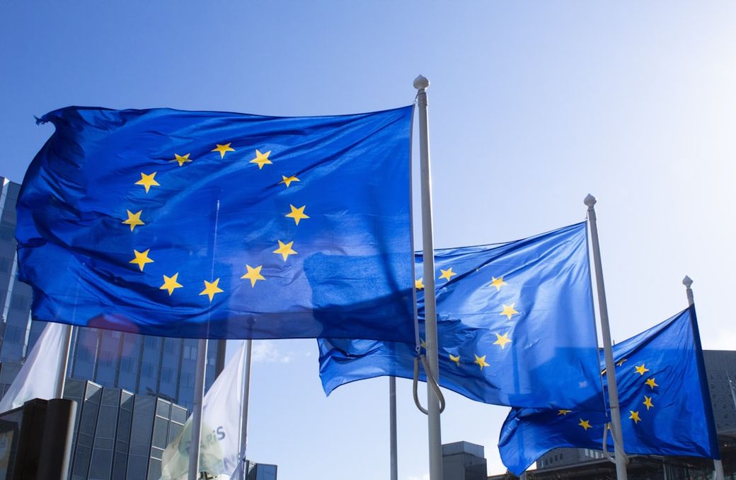 The image features multiple European Union flags waving in the breeze against a clear blue sky. The flags are prominently displayed, highlighting their stars and colors. — Credit: Photo by ALEXANDRE LALLEMAND on Unsplash