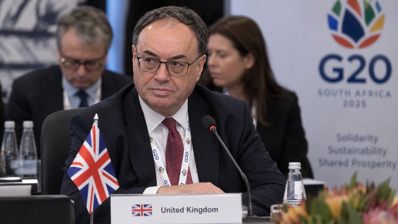 The image shows Andrew Bailey, the Governor of the Bank of England, seated at a conference table during the G20 summit in South Africa. He is engaged in discussions, with a United Kingdom flag and a placard identifying him, while other delegates are visible in the background.