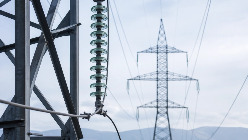 The image shows high-voltage power lines and a transmission tower against a cloudy sky. The foreground features a close-up of an insulator on a power line, emphasizing the electrical infrastructure.