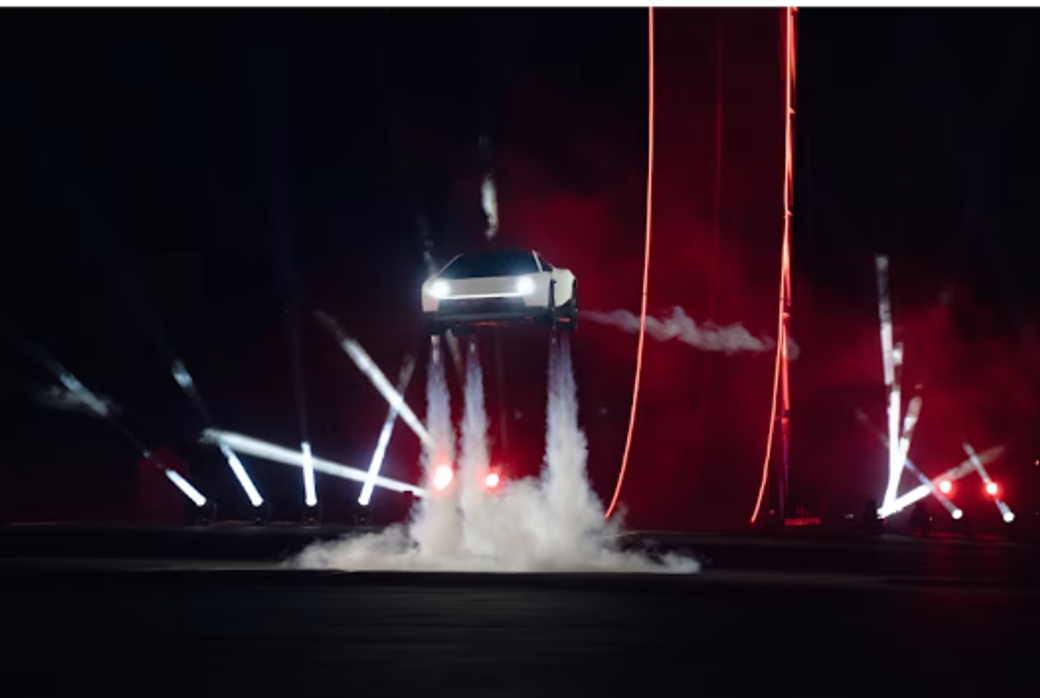 An electric vehicle performing a dramatic jump against a dark backdrop illuminated by bright stage lights. The vehicle appears to be generating smoke as it takes off, emphasizing a high-tech and thrilling atmosphere.