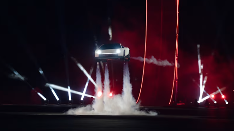 An electric vehicle performing a dramatic jump against a dark backdrop illuminated by bright stage lights. The vehicle appears to be generating smoke as it takes off, emphasizing a high-tech and thrilling atmosphere.