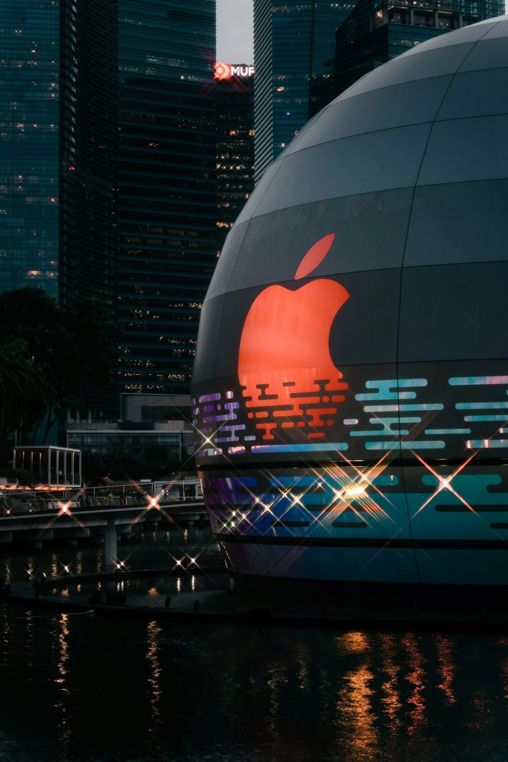 The image showcases a large illuminated Apple logo on a modern building exterior. The surrounding area features urban architecture with tall buildings in the background. — Credit: Photo by Keming Tan / Unsplash