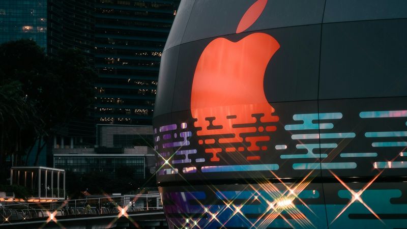 The image showcases a large illuminated Apple logo on a modern building exterior. The surrounding area features urban architecture with tall buildings in the background.
