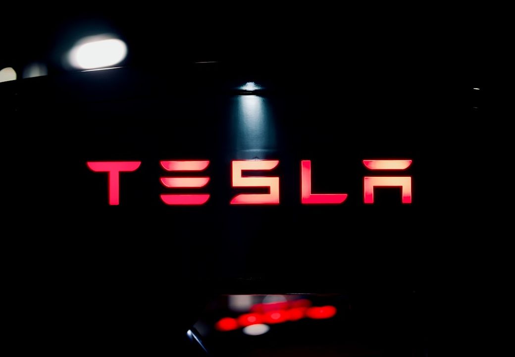 The image features the illuminated logo of Tesla, prominently displayed against a dark background. The sleek design highlights the brand's modern and innovative identity. — Credit: Photo by Prometheus 🔥 on Unsplash