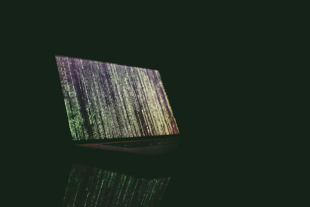 The image features a laptop displaying streams of colorful data code on its screen, set against a dark backdrop. The reflective surface beneath the laptop enhances the visual impact of the glowing text. — Credit: Photo by Markus Spiske / Unsplash