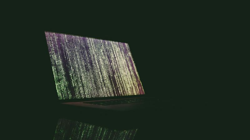 The image features a laptop displaying streams of colorful data code on its screen, set against a dark backdrop. The reflective surface beneath the laptop enhances the visual impact of the glowing text.