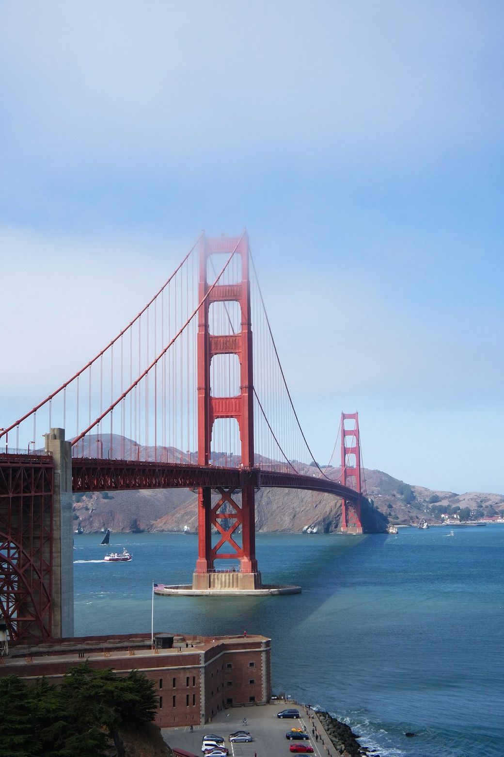 A scenic view of the Golden Gate Bridge partially shrouded in fog, spanning across the water with a backdrop of a hilly landscape. The bridge's iconic red-orange color stands out against the blue sea and cloudy sky. — Credit: Photo by Michaela on Unsplash
