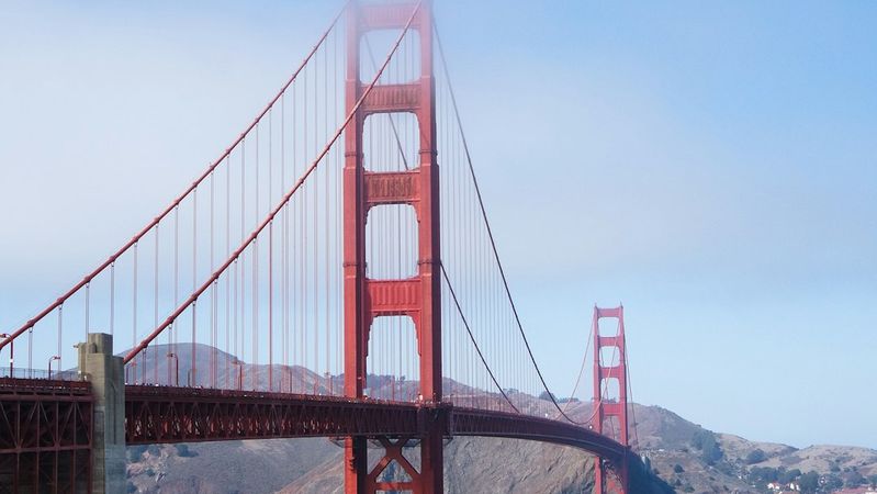 A scenic view of the Golden Gate Bridge partially shrouded in fog, spanning across the water with a backdrop of a hilly landscape. The bridge's iconic red-orange color stands out against the blue sea and cloudy sky.