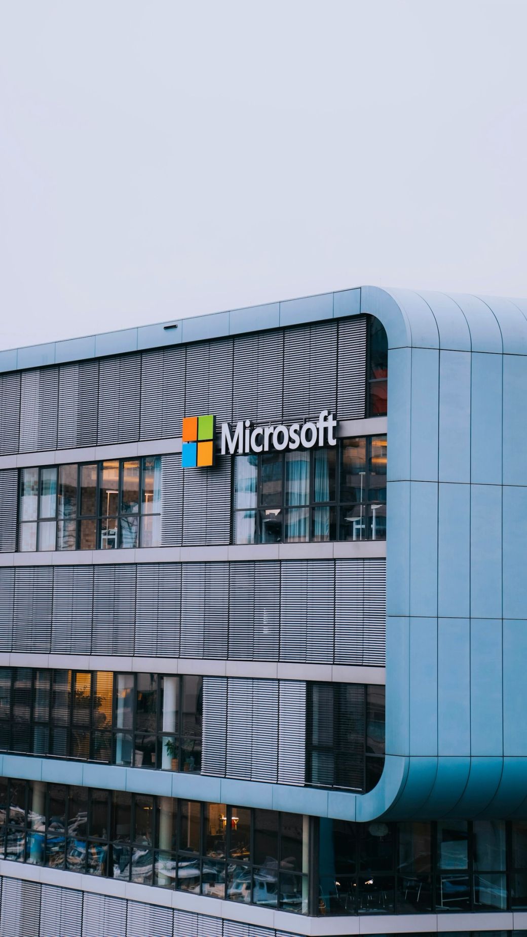 The image features the exterior of a modern building with large windows and a prominent Microsoft logo displayed on the wall. The architecture showcases contemporary design elements with horizontal lines. — Credit: Photo by ‪Salah Darwish / Unsplash