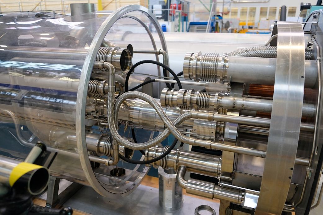 The image showcases a close-up view of a complex metallic apparatus, featuring numerous pipes and connectors within a transparent enclosure. The setting appears to be a high-tech engineering or research facility. — Credit: Photo by Brandon Style on Unsplash