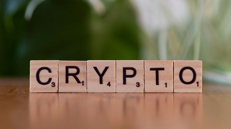 The image features wooden letter tiles arranged to spell the word 'CRYPTO.' The background is blurred, suggesting a natural setting.