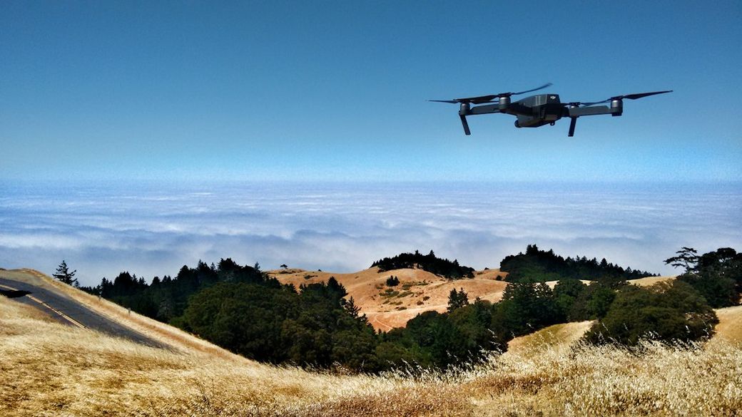 A drone is flying over a picturesque landscape that features rolling hills and a view of a foggy horizon. The scene captures the contrast between the lush green vegetation and the golden dry grass. — Credit: Photo by Ian Usher on Unsplash