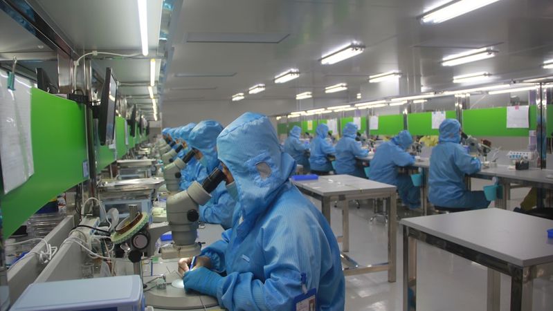 The image depicts a cleanroom environment where multiple researchers, dressed in blue protective suits, are engaged in scientific work. Each researcher is using a microscope at their workstation, demonstrating a focus on precision and safety in their tasks.