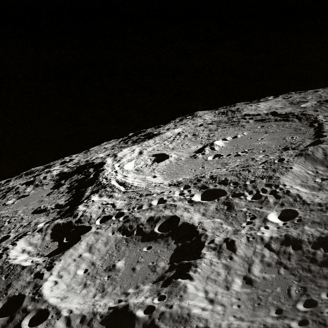 The image depicts a close-up view of the moon's surface, showcasing its craters and rugged terrain. The contrast between light and shadow emphasizes the texture of the lunar landscape.