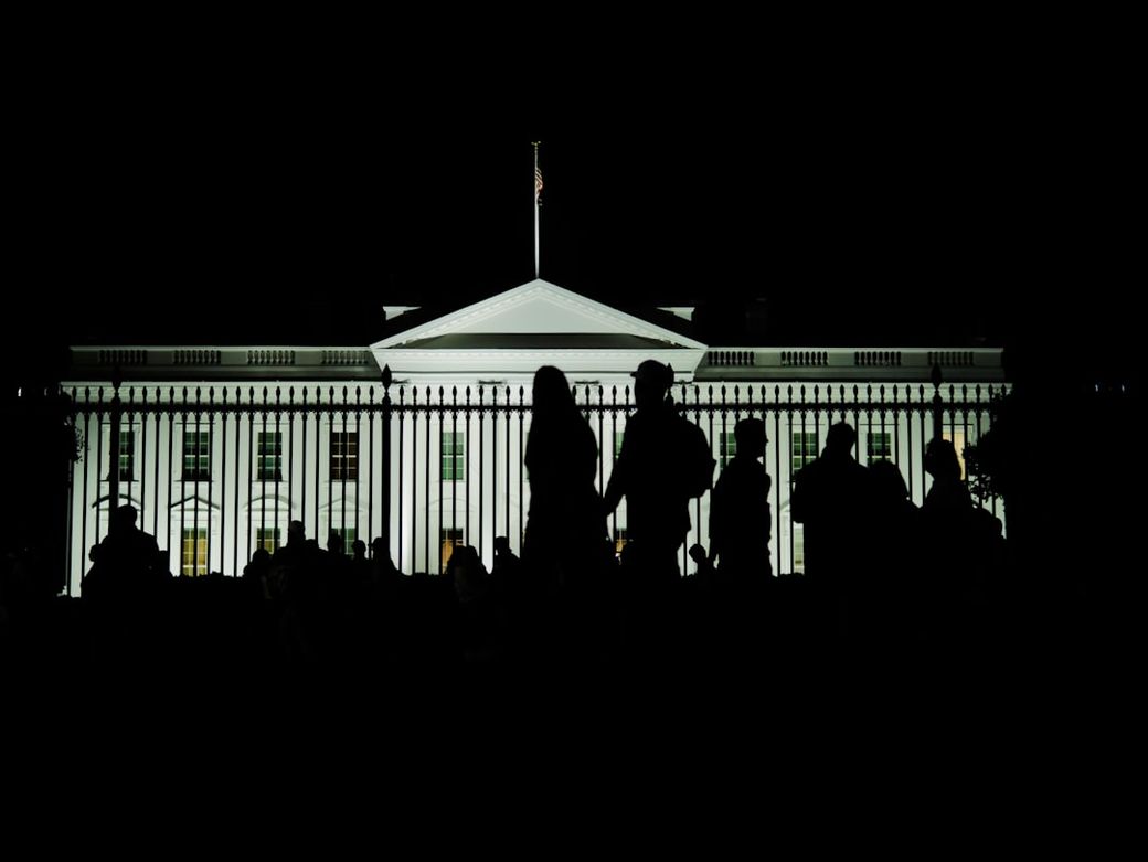 The image shows the silhouette of a crowd in front of the White House, illuminated at night. The iconic architecture of the White House is clearly visible, surrounded by a fence. — Credit: Photo by Jonathan Ardila on Unsplash
