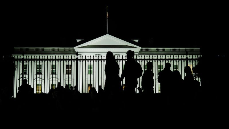 The image shows the silhouette of a crowd in front of the White House, illuminated at night. The iconic architecture of the White House is clearly visible, surrounded by a fence.