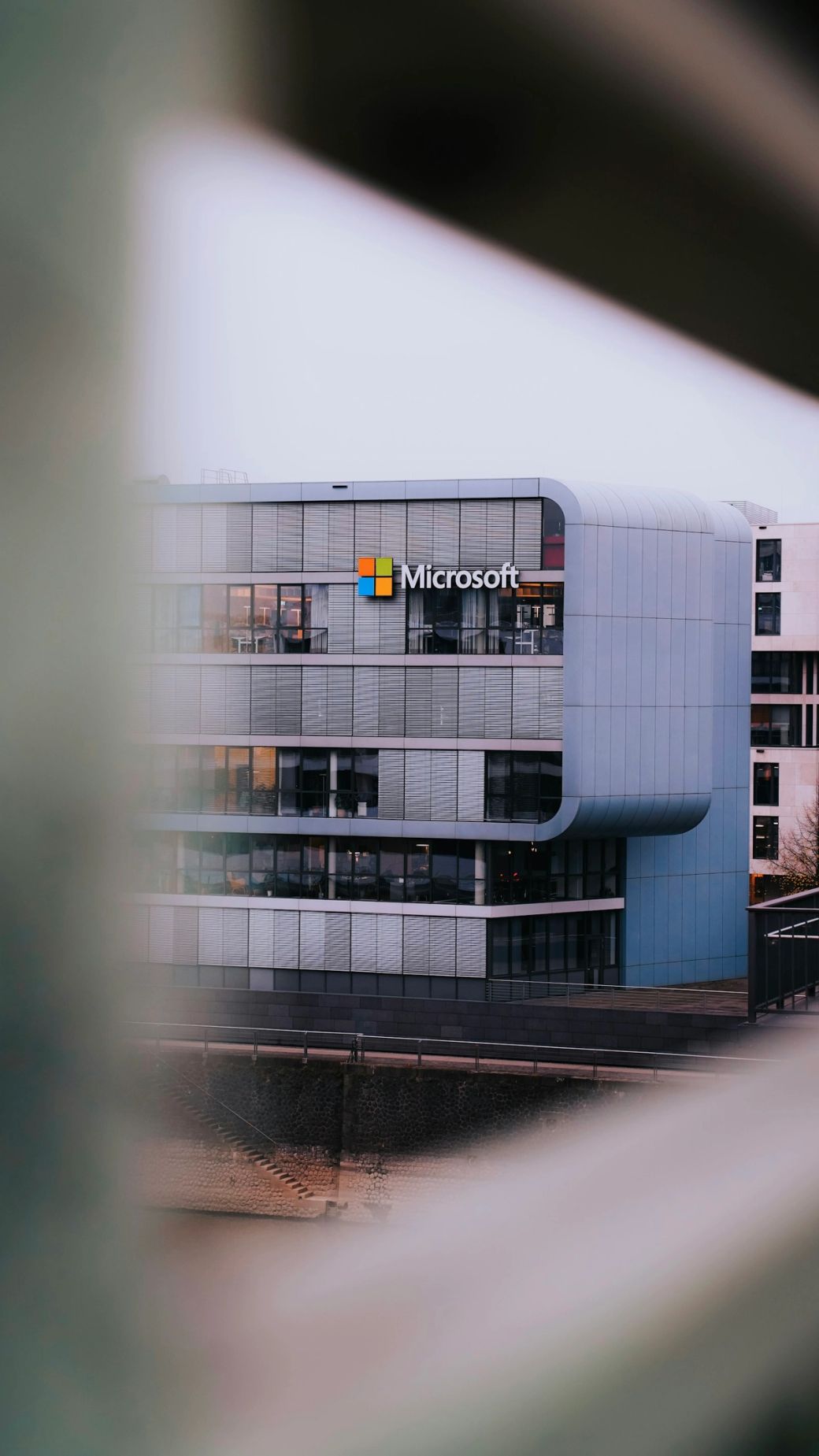 The image shows the exterior of a modern Microsoft office building, highlighting the company's logo prominently displayed on the facade. The architectural design features a sleek, contemporary look with large windows. — Credit: Photo by ‪Salah Darwish / Unsplash