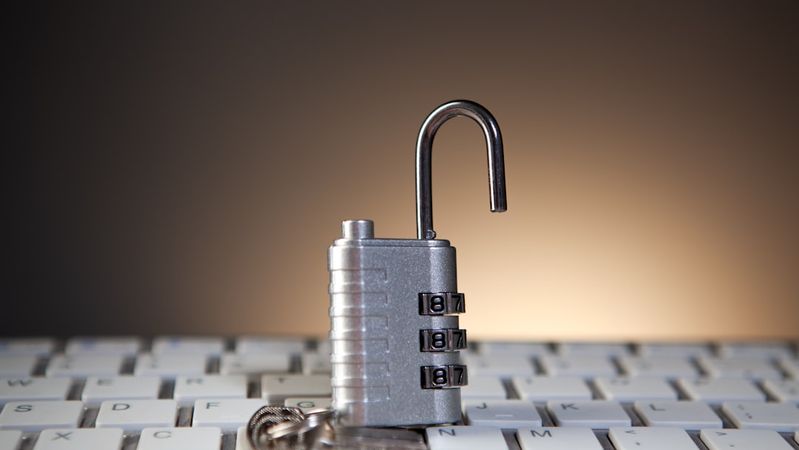 The image features a silver combination lock placed on a computer keyboard, with the lock appearing partially open. The blurred background suggests a focused setting on digital security themes.