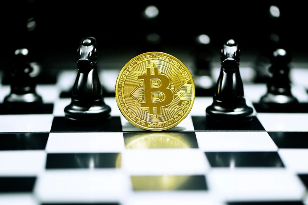 The image features a large golden Bitcoin symbol prominently displayed on a chessboard, with black chess pieces in the background. This composition juxtaposes the concepts of cryptocurrency and strategy. — Credit: Photo by Kanchanara / Unsplash