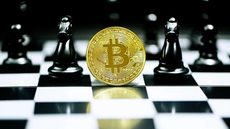 The image features a large golden Bitcoin symbol prominently displayed on a chessboard, with black chess pieces in the background. This composition juxtaposes the concepts of cryptocurrency and strategy.