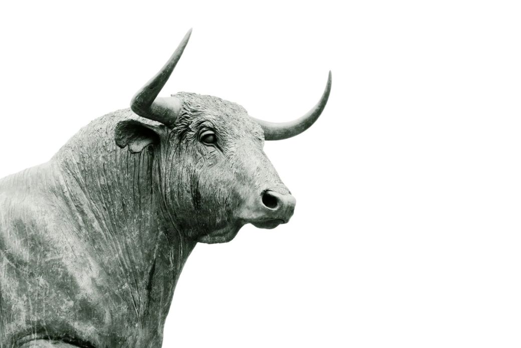 The image features a close-up of a bull statue, showcasing detailed textures and features of the animal. The bull is depicted in a monochromatic style, emphasizing its strength and prominence. — Credit: Photo by Hans Eiskonen / Unsplash