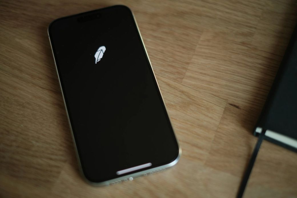 The image shows a smartphone displaying the logo of a trading application on a wooden desk. Next to the phone, there is a notebook, suggesting a setting for personal finance management. — Credit: Photo by appshunter.io / Unsplash