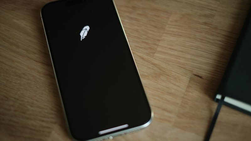 The image shows a smartphone displaying the logo of a trading application on a wooden desk. Next to the phone, there is a notebook, suggesting a setting for personal finance management.