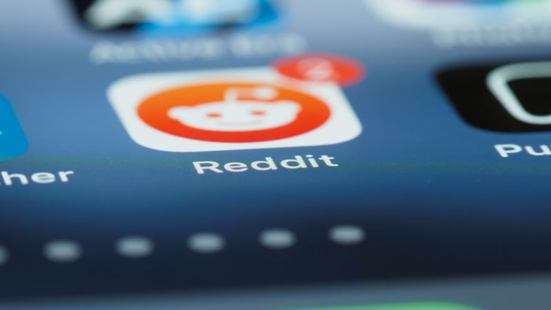 The image displays a close-up view of a smartphone screen showcasing various app icons, prominently featuring the Reddit app with a notification badge. The background is blurred, emphasizing the app's icon.