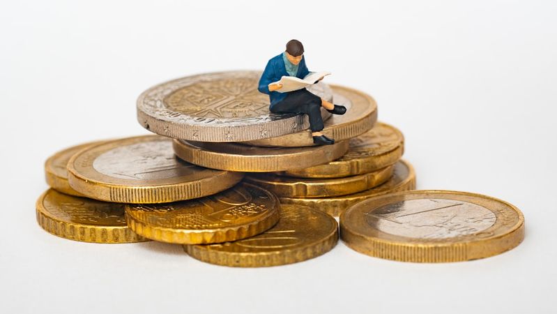 The image features a miniature figurine of a person reading a book while sitting on top of oversized coins. This composition illustrates the concept of finance and investment through a creative, symbolic representation.