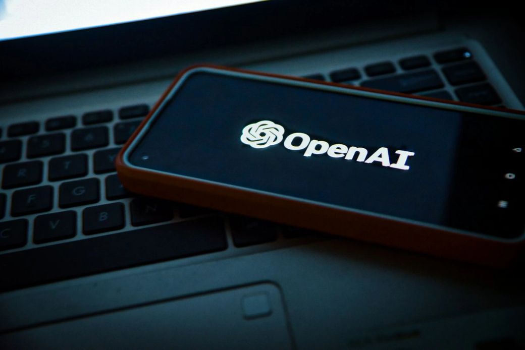 An illuminated smartphone displaying the OpenAI logo is placed on a laptop keyboard. The scene is dimly lit, highlighting the contrast between the phone's screen and the keyboard. — Credit: Photo by Levart_Photographer / Unsplash