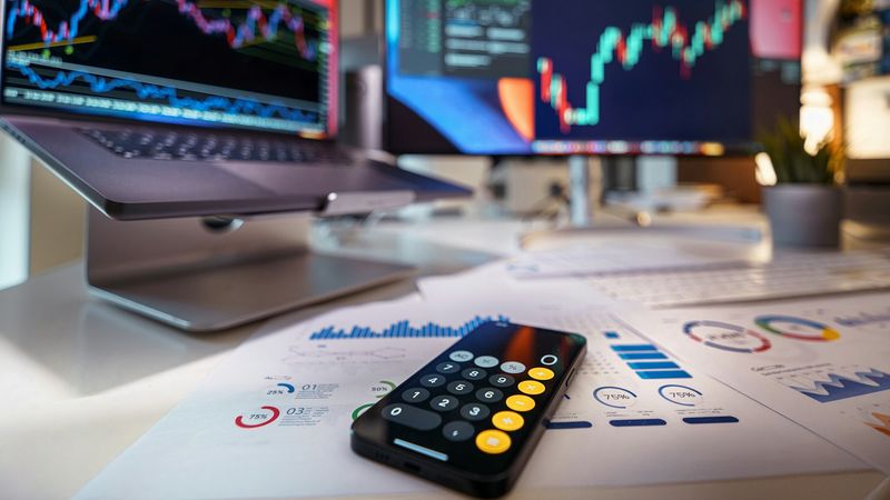 The image displays a close-up view of a trading workspace featuring multiple monitors displaying stock market graphs and data analysis. A calculator and printed reports are positioned on a desk, suggesting active financial analysis and trading activities.