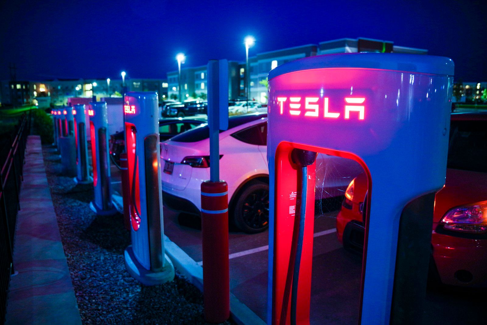 A Tesla charging station at night featuring multiple electric vehicles parked nearby. The charging units are lit with red illumination, showcasing the Tesla logo prominently.