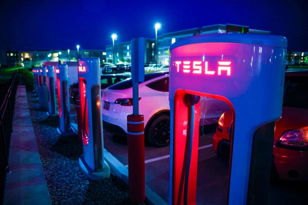 A Tesla charging station at night featuring multiple electric vehicles parked nearby. The charging units are lit with red illumination, showcasing the Tesla logo prominently. — Credit: Photo by Stephen Mease / Unsplash