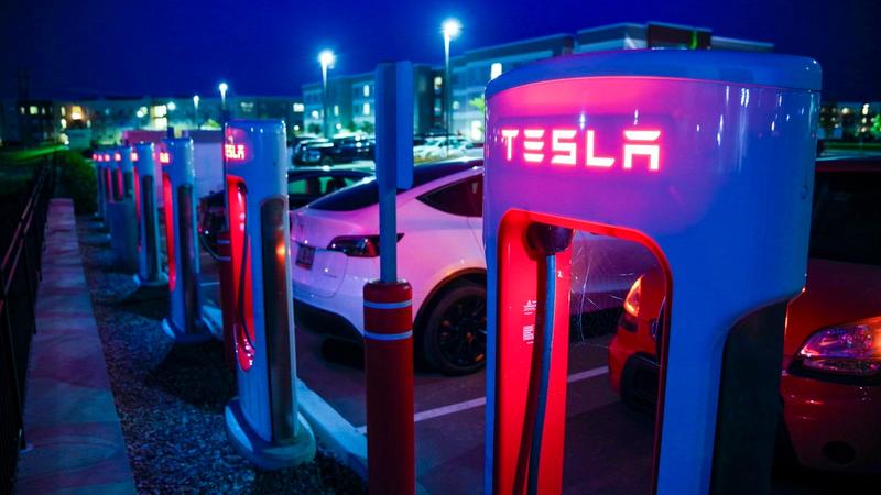 A Tesla charging station at night featuring multiple electric vehicles parked nearby. The charging units are lit with red illumination, showcasing the Tesla logo prominently.