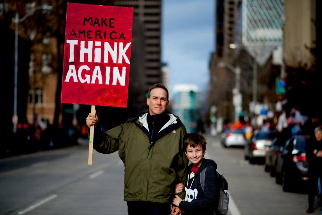 The image depicts a man and a boy standing on a city street, holding a large red sign that reads 'MAKE AMERICA THINK AGAIN.' The background shows a crowd of people and urban buildings, suggesting a public demonstration or rally. — Credit: Photo by Jose M / Unsplash