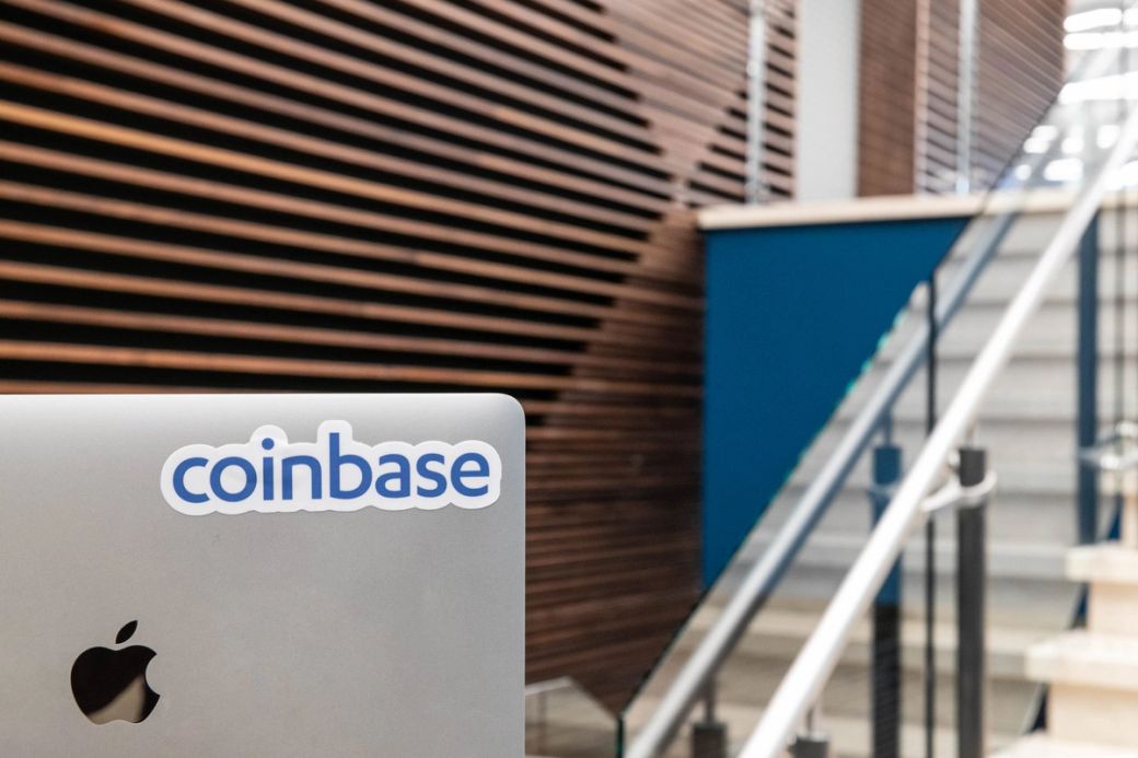 The image features a close-up view of a laptop with a sticker displaying the logo of Coinbase, set against a backdrop of a modern interior with wooden paneling and a staircase.
