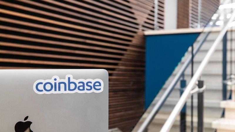The image features a close-up view of a laptop with a sticker displaying the logo of Coinbase, set against a backdrop of a modern interior with wooden paneling and a staircase.