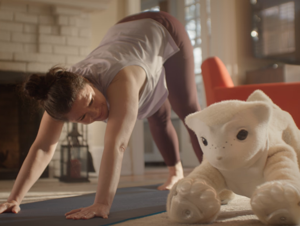 A woman practices yoga at home, performing a downward-facing dog pose on a yoga mat. A white plush toy resembling a cat is positioned beside her on the mat, adding a playful touch to the scene.