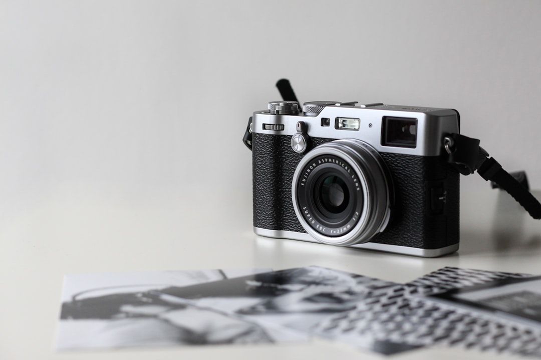 The image features a vintage-style camera placed on a flat surface alongside printed photographs. The camera is well-detailed, showcasing its lens and controls, while the photos appear to depict black-and-white imagery, suggesting a focus on photography as an art form.