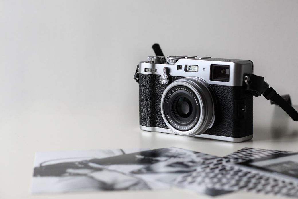 The image features a vintage-style camera placed on a flat surface alongside printed photographs. The camera is well-detailed, showcasing its lens and controls, while the photos appear to depict black-and-white imagery, suggesting a focus on photography as an art form. — Credit: Photo by NordWood Themes on Unsplash